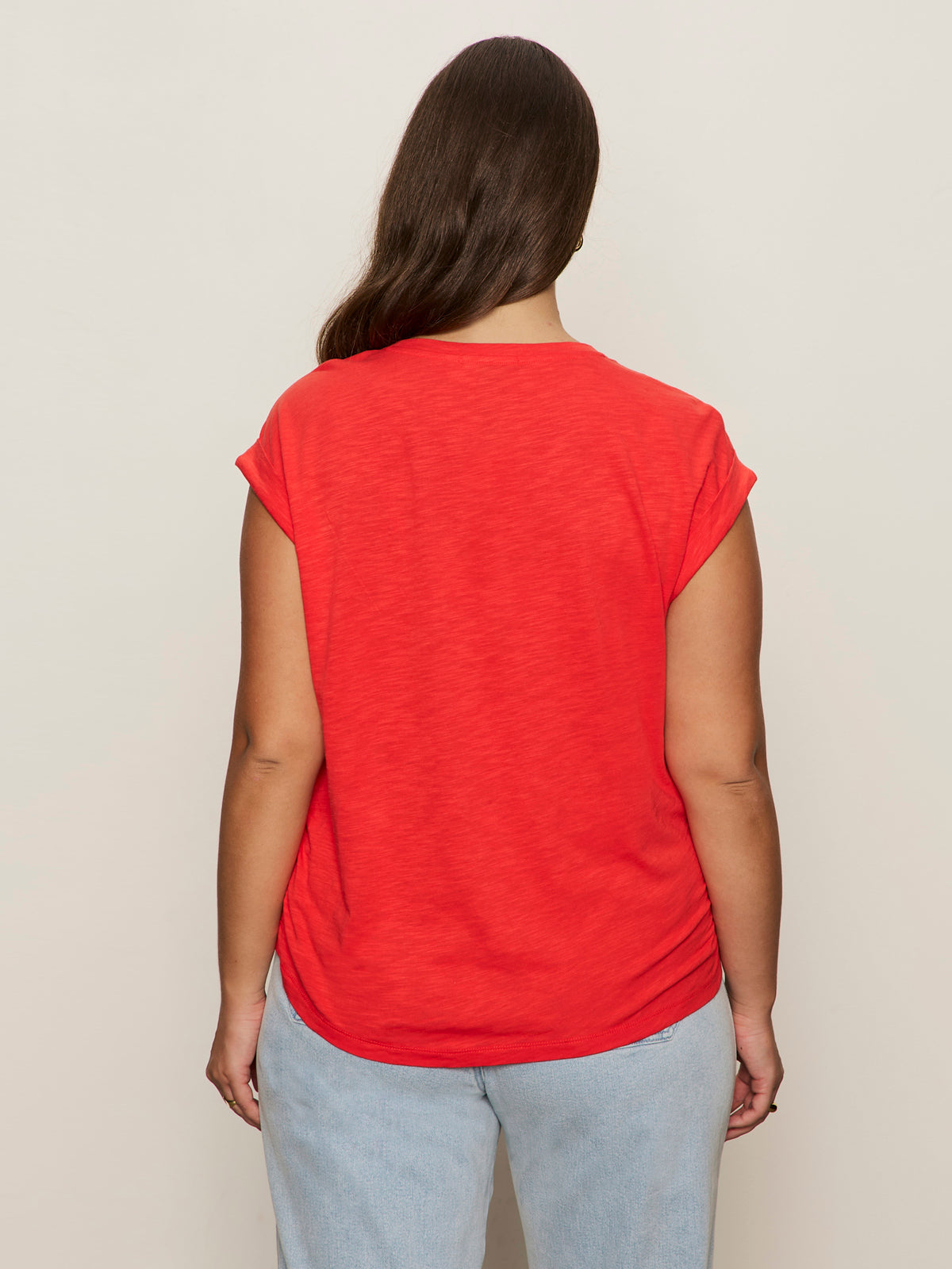 A person with long brown hair stands facing away, wearing the Sanctuary Clothing knotted muscle tee in cayenne (extended sizing) and light blue jeans, set against a plain beige background.
