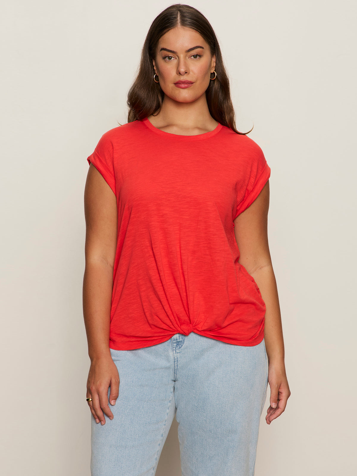 A woman with long brown hair wears the Sanctuary Clothing knotted muscle tee in cayenne extended sizing and light blue jeans, standing against a plain light background.