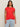 A woman with long brown hair wears the Sanctuary Clothing knotted muscle tee in cayenne extended sizing and light blue jeans, standing against a plain light background.