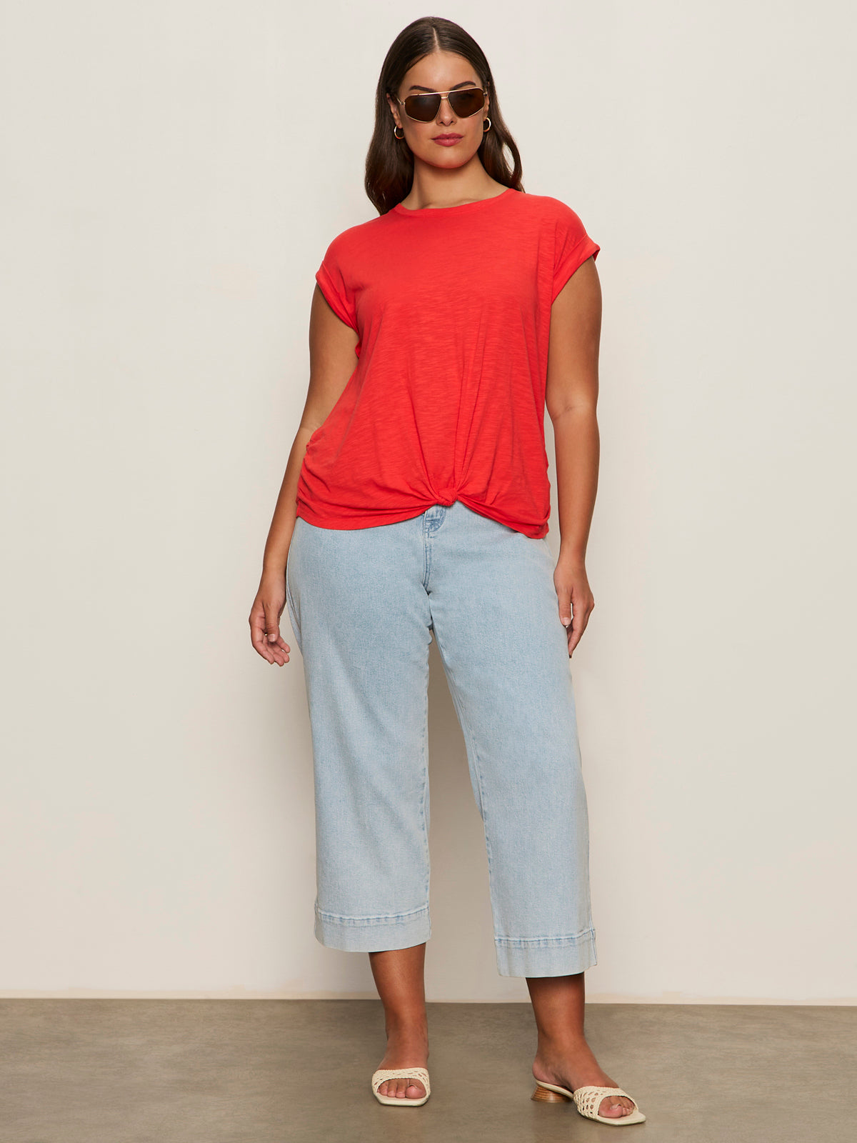 A woman wearing Sanctuary Clothing's knotted muscle tee in cayenne, light blue wide-leg jeans, sunglasses, and beige slide sandals stands against a plain beige background.