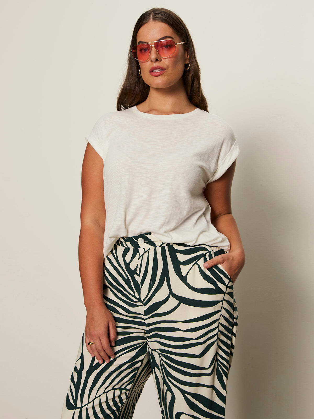 A woman with long brown hair wears pink sunglasses and the Sanctuary Clothing knotted muscle tee in white (extended sizing), paired with high-waisted green and white leaf print pants, standing against a plain light background with one hand in her pocket.