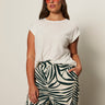 A woman with long brown hair wears pink sunglasses and the Sanctuary Clothing knotted muscle tee in white (extended sizing), paired with high-waisted green and white leaf print pants, standing against a plain light background with one hand in her pocket.