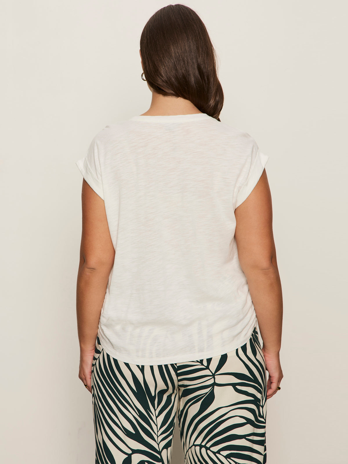 A woman, seen from behind with long side-parted hair, wears the Sanctuary Clothing knotted muscle tee in white (extended sizing) and high-waisted black and white tropical leaf pants against a plain light background.