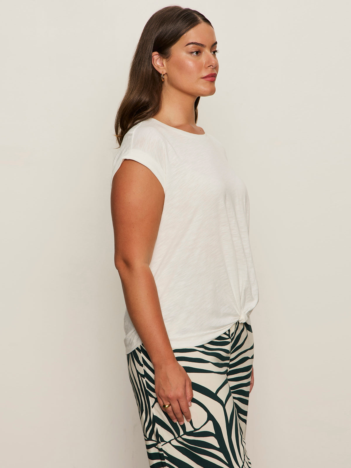 A woman stands in profile against a plain background, wearing the Sanctuary Clothing knotted muscle tee in white (extended sizing) paired with high-waisted zebra print pants. She has long brown hair and minimal accessories.