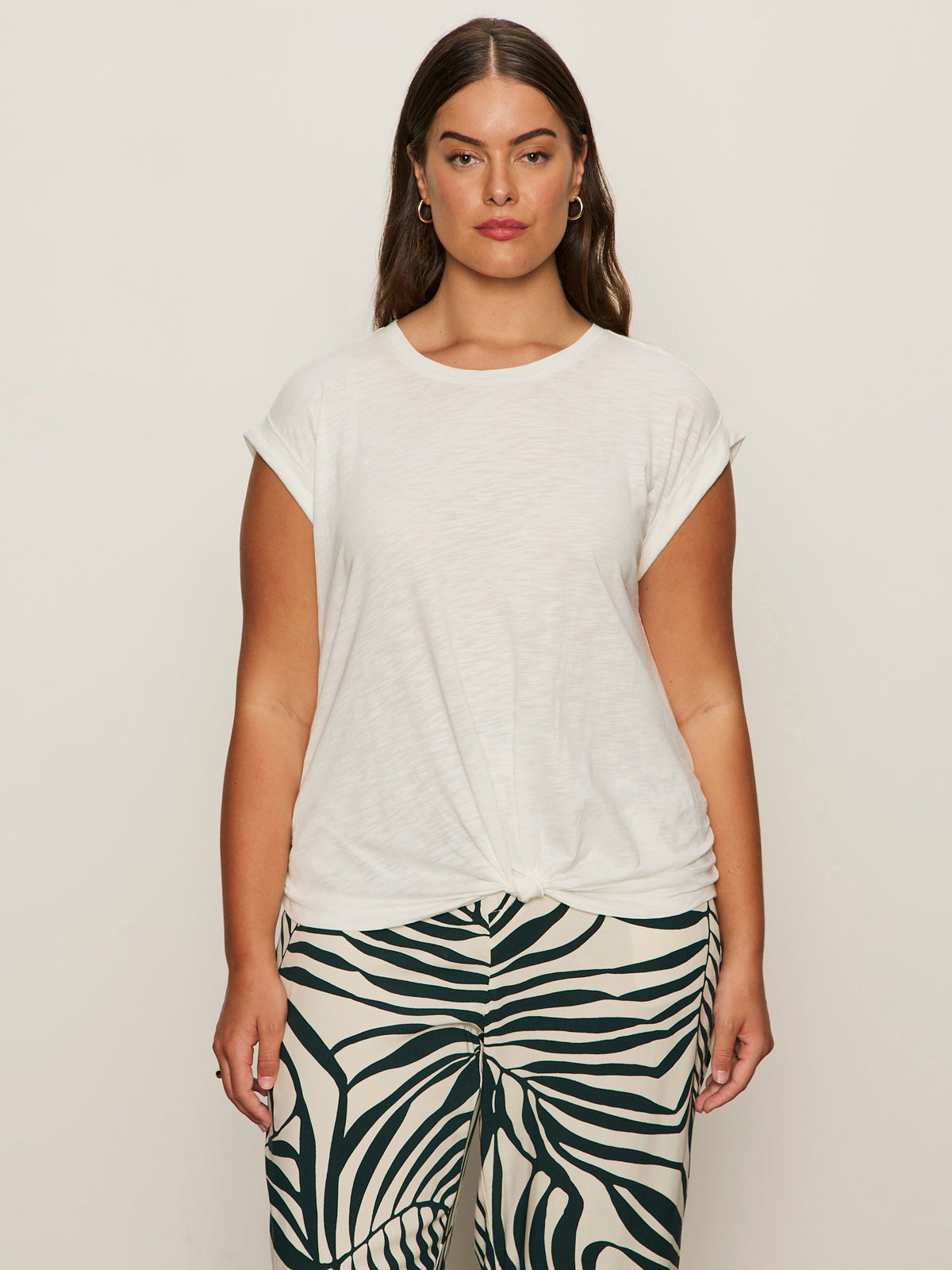 A woman with long brown hair faces forward against a plain background, wearing Sanctuary Clothing's knotted muscle tee in white (extended sizing) and high-waisted black and white zebra print pants.