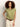 A woman with curly hair models the Sanctuary Clothing knotted muscle tee bamboo in light green, paired with beige pants and a brown belt, posing against a plain light background with one hand on her hip.