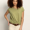 A woman with curly hair models the Sanctuary Clothing knotted muscle tee bamboo in light green, paired with beige pants and a brown belt, posing against a plain light background with one hand on her hip.