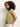 A woman with curly brown hair stands sideways in a light green Sanctuary Clothing "knotted muscle tee bamboo" and cream pants with a brown belt, set against a plain light background.