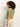 A person with curly brown hair stands with their back to the camera, wearing a light green Sanctuary Clothing knotted muscle tee bamboo and off-white pants against a plain light background.