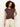 A woman with curly brown hair wears the Sanctuary Clothing knotted muscle tee mesa—a loose, short-sleeved brown top with a front knot—paired with cream-colored pants, standing against a plain light background.