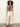 A woman with curly hair stands against a plain light wall wearing the Sanctuary Clothing knotted muscle tee in mesa palm, cream wide-leg pants, and brown sandals. She has a relaxed expression and her arms rest by her sides.