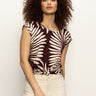 A woman with curly hair wears the Sanctuary Clothing knotted muscle tee in mesa palm print, paired with a light skirt, against a plain light background.