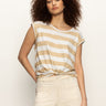 A woman with curly hair wears the Sanctuary Clothing knotted muscle tee in white sandstone stripe and light beige pants, standing against a plain background with a relaxed expression.