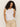 A person with long, curly hair wearing the "linen tank white" by Sanctuary Clothing and beige pants stands against a plain background, gazing directly at the camera.
