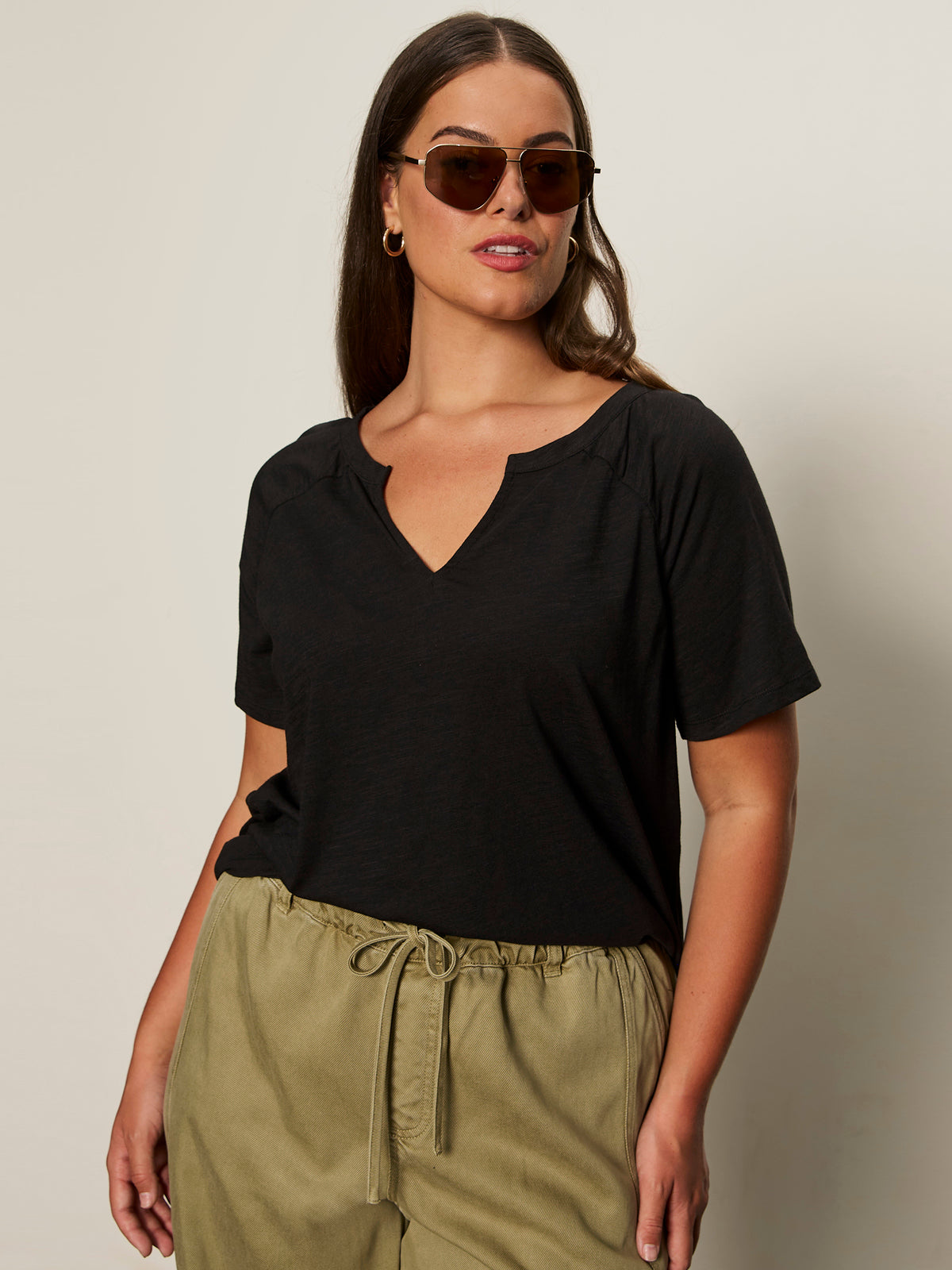 A woman with long brown hair wears sunglasses and the Sanctuary Clothing notch neck tee in black extended sizing, paired with light olive drawstring pants, standing against a plain light background.