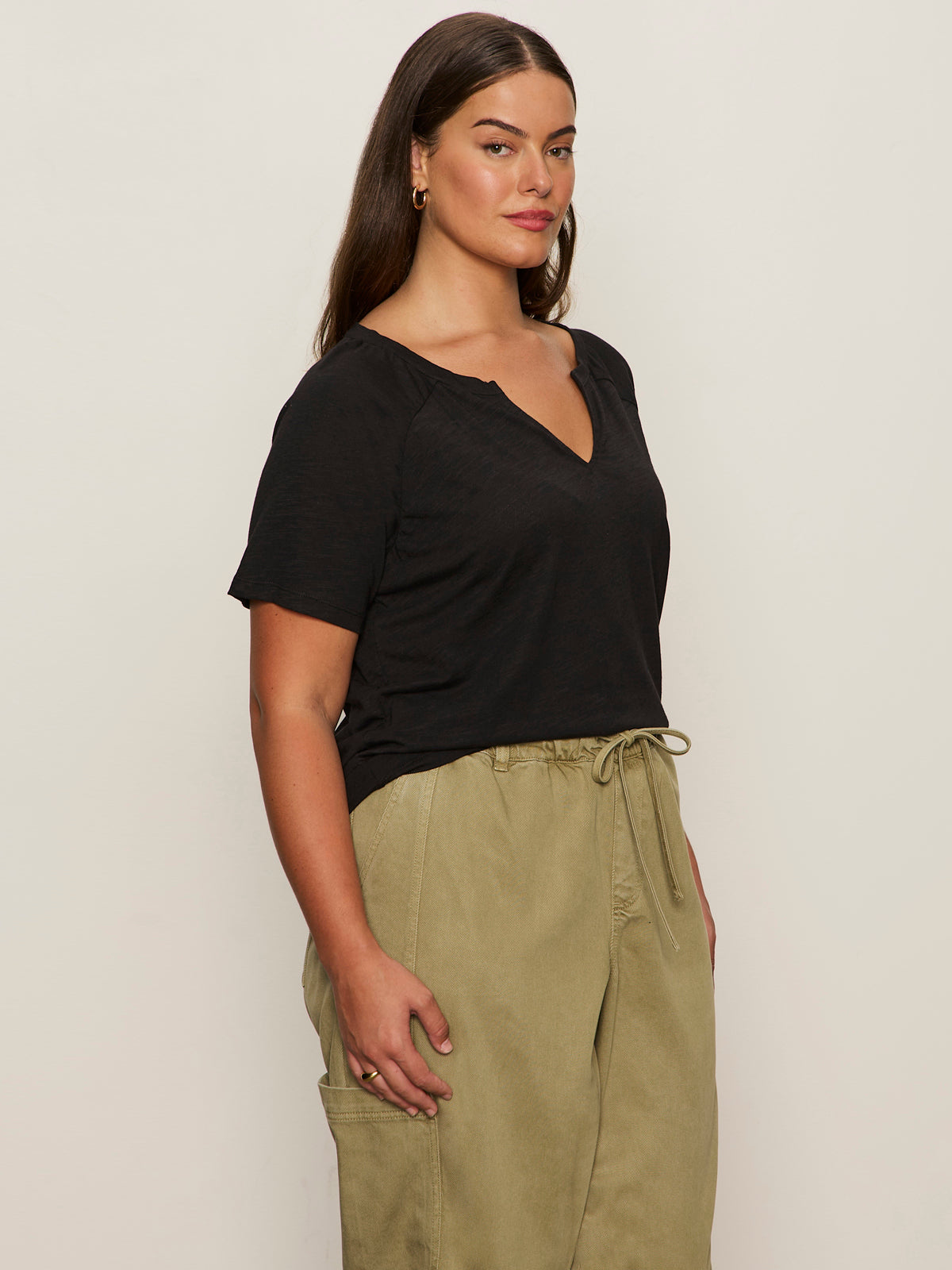A person with long brown hair wears the Sanctuary Clothing notch neck tee in black (extended sizing) and loose-fitting olive green drawstring pants, standing against a plain light background and looking slightly to the side.