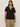 A woman with long brown hair wears the Sanctuary Clothing notch neck tee in black (extended sizing), paired with light olive green pants, standing against a plain, light-colored background.
