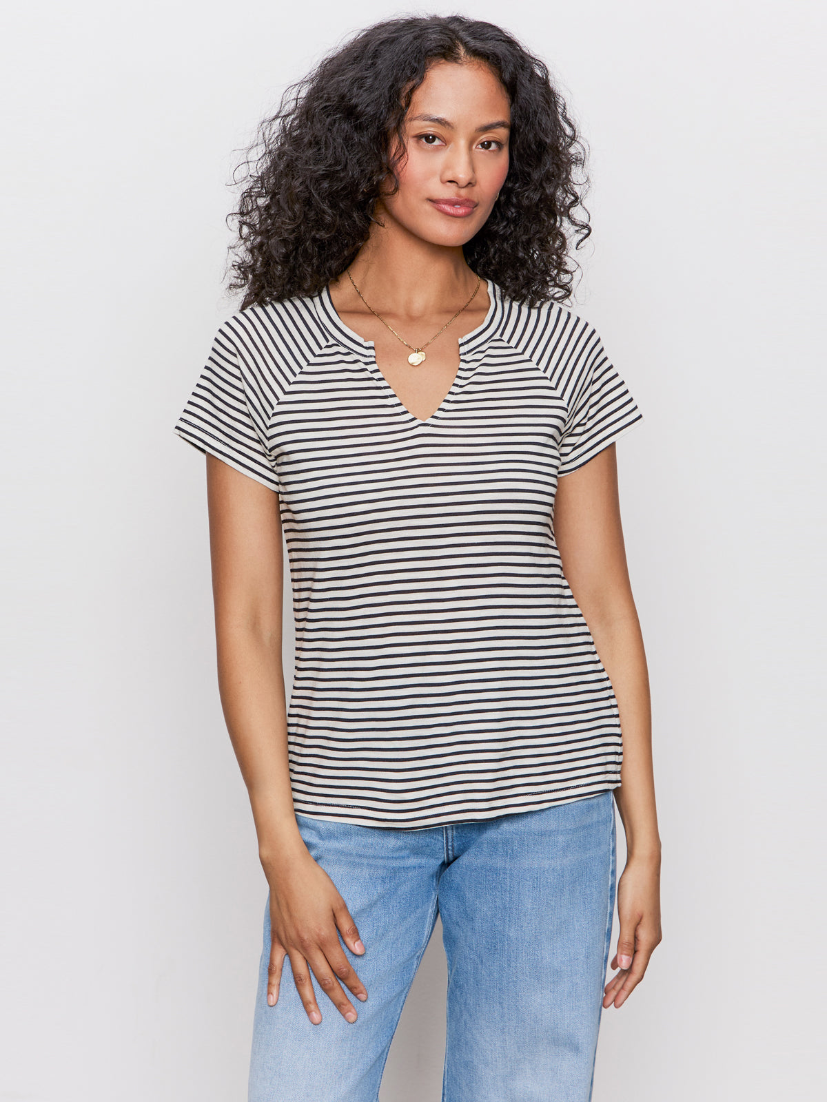 A woman with curly hair wears the Sanctuary Clothing notch neck tee in light oat black stripe, paired with light blue jeans, standing against a plain light background.