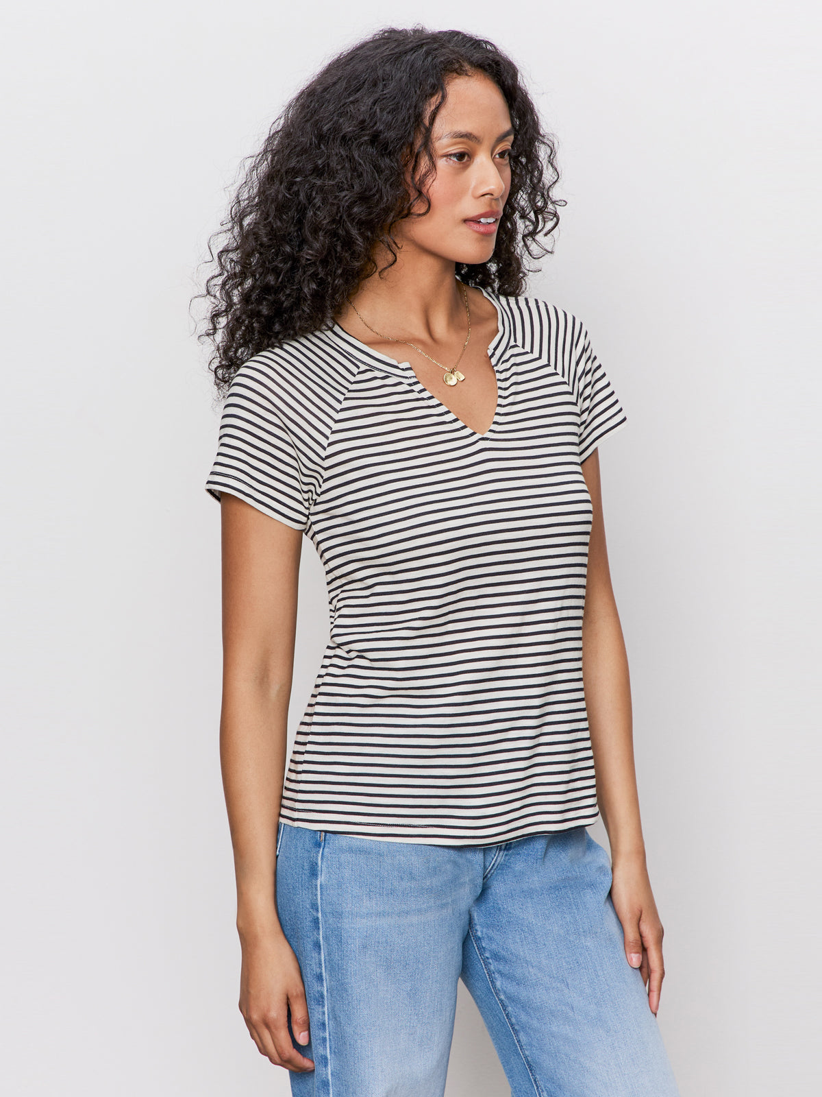 Woman with long curly hair wears the Sanctuary Clothing notch neck tee in light oat black stripe 2 and light blue jeans, standing against a plain light background, looking to the side.
