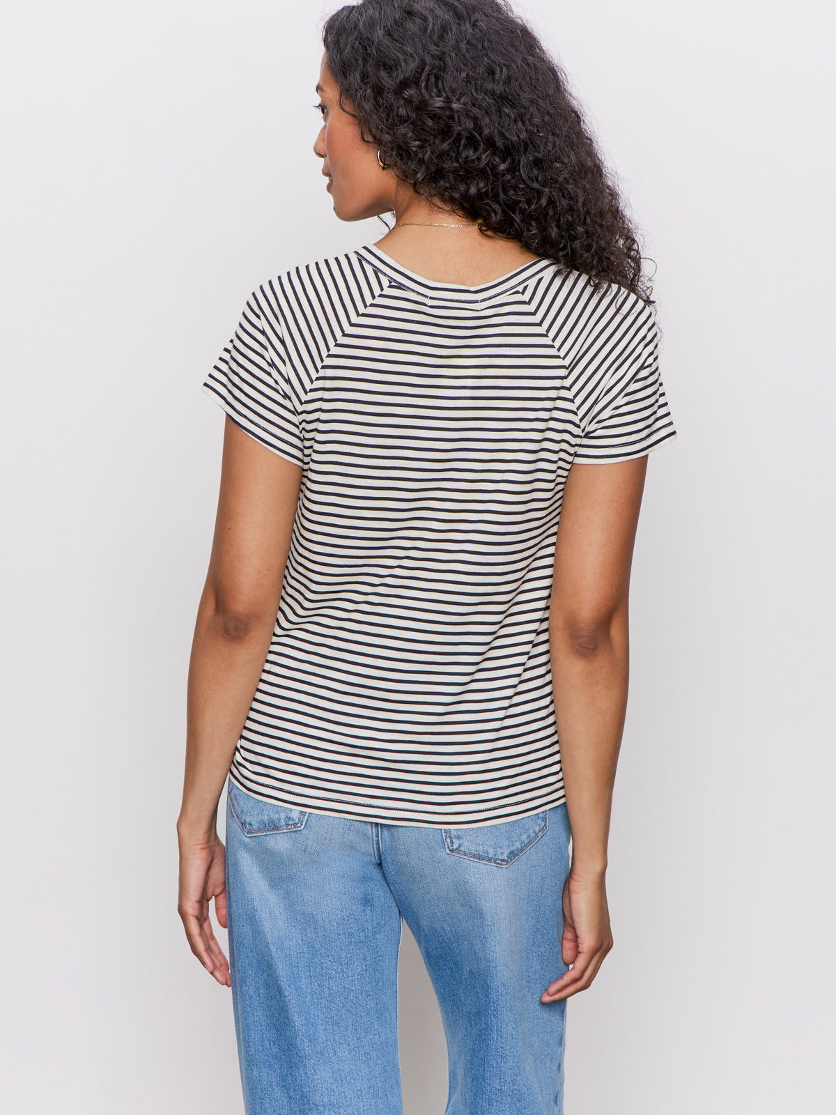 A woman with curly hair is shown from the back wearing the Sanctuary Clothing notch neck tee light oat black stripe 2 and light blue jeans, standing against a plain white background.