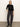 A woman stands against a plain light background wearing the Sanctuary Clothing relaxed sparkle popover black sweater, dark gray cargo pants, and black heeled ankle boots, left hand in her pocket and a neutral expression.