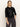 A woman with long, straight blonde hair stands against a plain light background wearing the Sanctuary Clothing relaxed sparkle popover in black and dark cargo pants, one hand in her pocket and a neutral expression.
