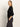 A woman with long blonde hair stands in profile, one hand in her pocket, wearing the Sanctuary Clothing relaxed sparkle popover black top and dark pants against a plain light background.