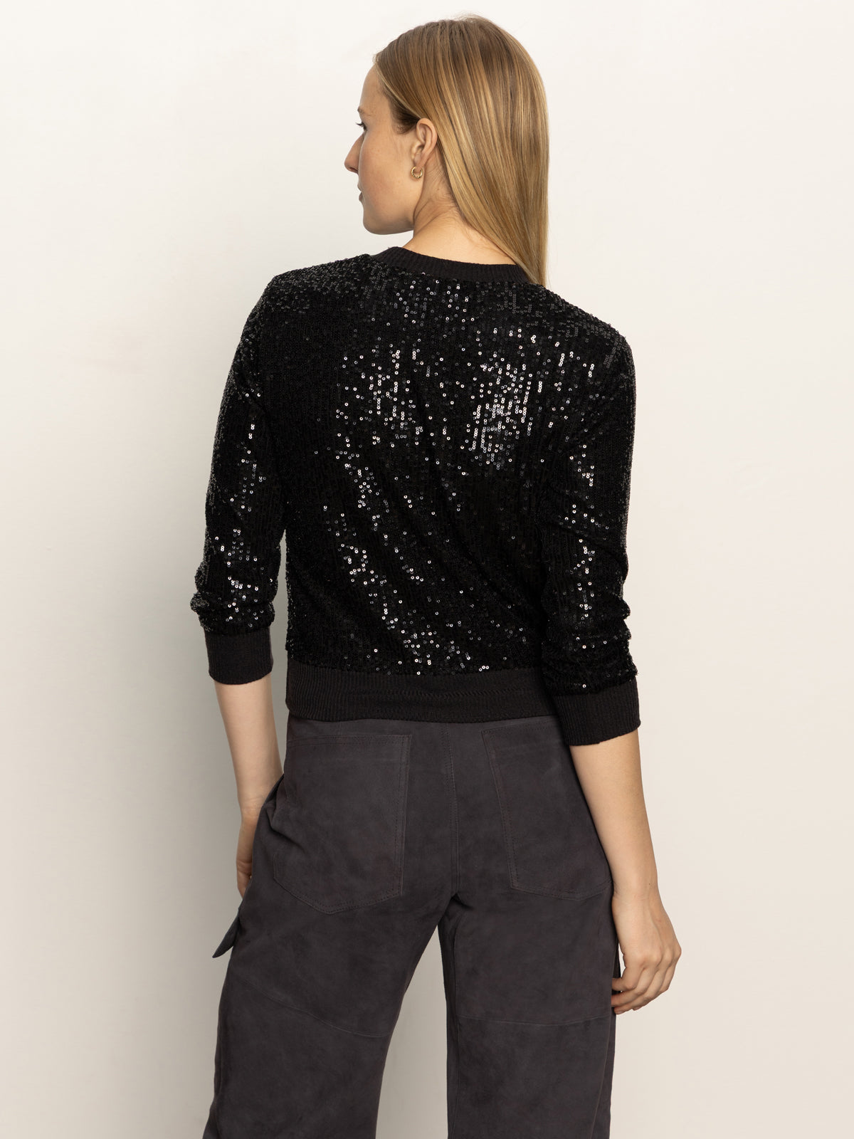 A woman with straight, light brown hair stands with her back to the camera, wearing the Sanctuary Clothing relaxed sparkle popover in black and dark high-waisted pants against a plain light background.