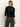 A woman with straight, light brown hair stands with her back to the camera, wearing the Sanctuary Clothing relaxed sparkle popover in black and dark high-waisted pants against a plain light background.
