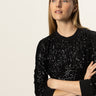 A woman with long blonde hair stands with her arms crossed, wearing the Sanctuary Clothing relaxed sparkle popover in black, against a plain light background.