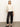 A woman stands against a plain light wall, wearing the Sanctuary Clothing relaxed sparkle popover chalk sweater with black satin pants and flats. One hand is in her pocket, and she looks relaxed.