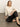 A woman with straight blonde hair sits against a plain background, wearing the Sanctuary Clothing relaxed sparkle popover in chalk, paired with black satin pants and black flats, looking confidently at the camera.