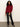 A woman with curly hair wears the Sanctuary Clothing change of pace top in crimson, black leather pants, and black pointed shoes, standing against a plain white wall and gray floor.