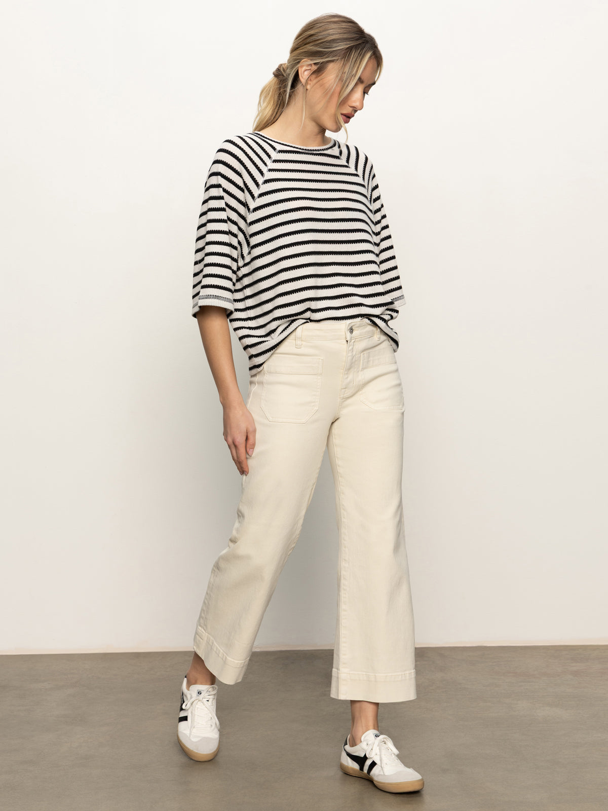 A woman stands against a plain background wearing the Sanctuary Clothing boxy raglan tee in black with light oat stripe, paired with off-white wide-leg pants and white sneakers with black accents, looking down with hands in her pockets.