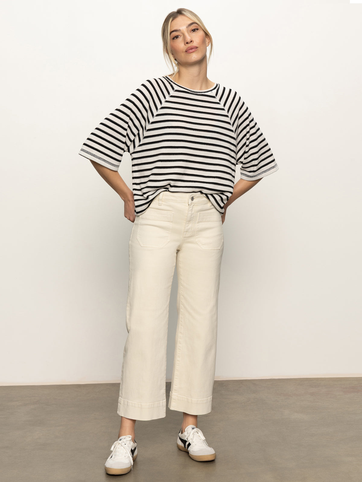 A woman stands against a plain background wearing the Sanctuary Clothing boxy raglan tee in black with light oat stripe. Her hands are behind her back and she looks confidently at the camera.
