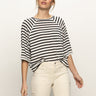 A woman stands against a white background wearing the Sanctuary Clothing boxy raglan tee in black with light oat stripe and high-waisted off-white pants, looking slightly to the side with a relaxed expression.