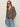 A woman wearing the Sanctuary Clothing dreamers top french kitty, sunglasses, and light blue jeans stands against a plain light background with one hand in her pocket.