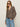 A woman wearing the Sanctuary Clothing dreamers top french kitty, sunglasses, and light blue jeans stands against a plain light background with one hand in her pocket.