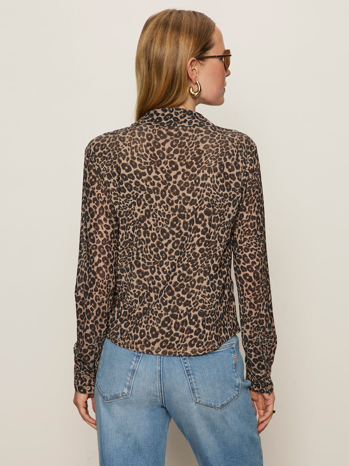 A person with light brown hair and sunglasses models the Sanctuary Clothing "dreamers top french kitty," a long-sleeve leopard print top, paired with blue jeans and hoop earrings, standing against a plain beige background.
