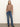 A woman stands against a plain wall wearing the Sanctuary Clothing long sleeve draped mock neck top in mocha, blue high-waisted jeans with front pockets, a brown belt, and black heeled boots. One hand is in her pocket as she looks at the camera.