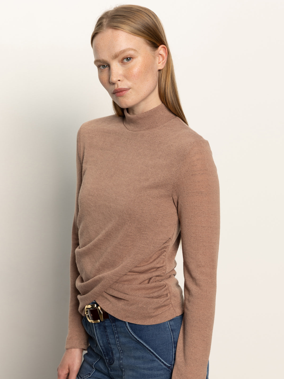 A woman with straight, light brown hair poses against a plain background, wearing the Sanctuary Clothing long sleeve draped mock neck top in mocha and blue jeans with a belt. She looks directly at the camera with a neutral expression.