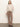 A woman with straight light brown hair and a neutral expression wears the Sanctuary Clothing long sleeve draped mock neck top in mother of pearl, paired with brown high-waisted bootcut pants and black heeled boots against a plain light wall.