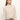 A woman with straight, light brown hair wears the Sanctuary Clothing long sleeve draped mock neck top in mother of pearl, paired with high-waisted brown pants, standing against a plain background and looking to her left.