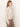 A woman with straight, light brown hair models the Sanctuary Clothing long sleeve draped mock neck top in Mother of Pearl, paired with high-waisted brown pants and photographed in profile against a plain white background.