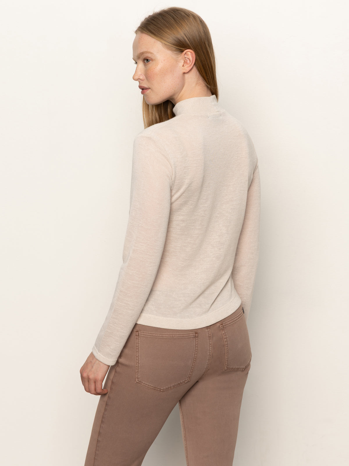 A woman with long straight hair, wearing the Sanctuary Clothing long sleeve draped mock neck top in mother of pearl, stands against a plain off-white background and looks over her shoulder.