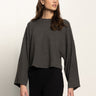 A woman with long brown hair wears the Sanctuary Clothing hearthside popover top in heather ink, a loose-fitting, wide-sleeve dark gray long-sleeve shirt, paired with black pants against a plain light background.