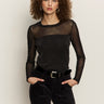 A woman with curly hair wears Sanctuary Clothing's sparkle mesh crew tee in black, paired with black pants and a silver-buckled belt, standing against a plain light background with her hands in her pockets.