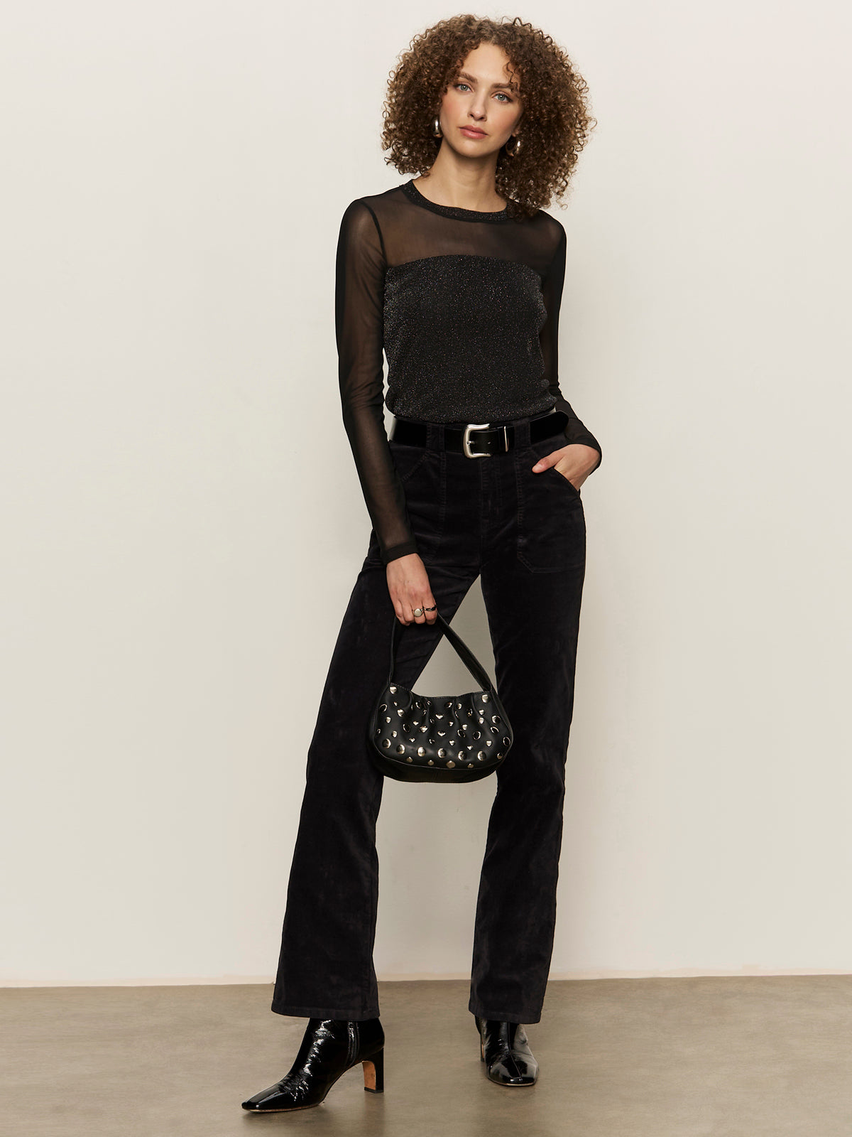 A woman with curly hair wears a sheer black long-sleeve top, black pants, and black boots. She holds a small black purse with silver studs and stands against a plain light-colored wall.