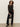 A woman with curly hair wears a sheer black long-sleeve top, black pants, and black boots. She holds a small black purse with silver studs and stands against a plain light-colored wall.
