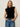 A woman with long blonde hair wears the Sanctuary Clothing pleated sleeveless top in black and blue jeans, accessorized with a black belt, standing against a neutral background with a relaxed expression.